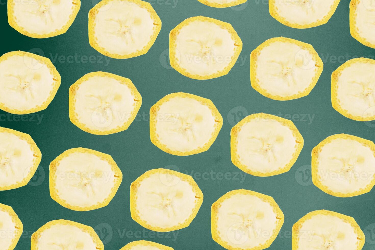 Round cut banana slices form a repeating grid pattern against a dark green background surface. photo