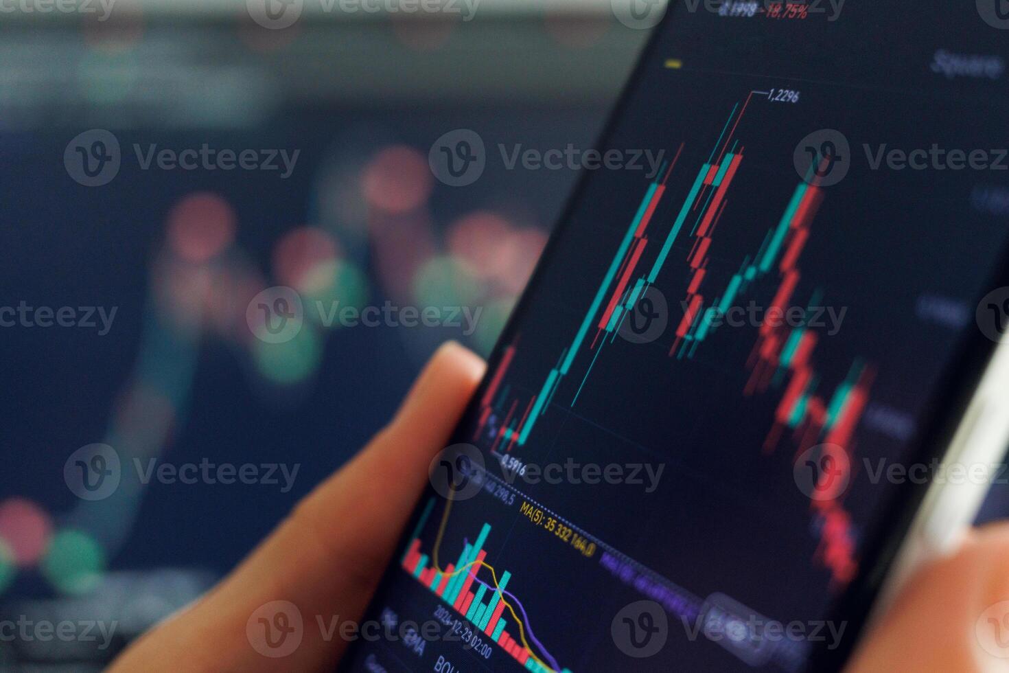 A hand holds a smartphone showing a fluctuating stock chart with red and green candlesticks. Another blurred screen displays similar data in the background. photo