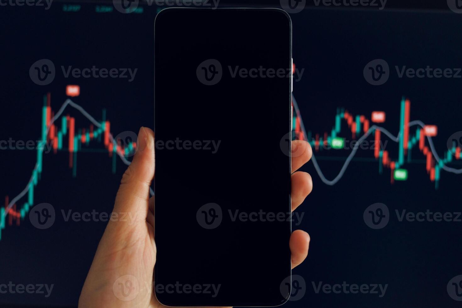 A hand holds a smartphone displaying a fluctuating stock chart with red and green candlesticks. Another blurred screen displays similar data in the background. photo