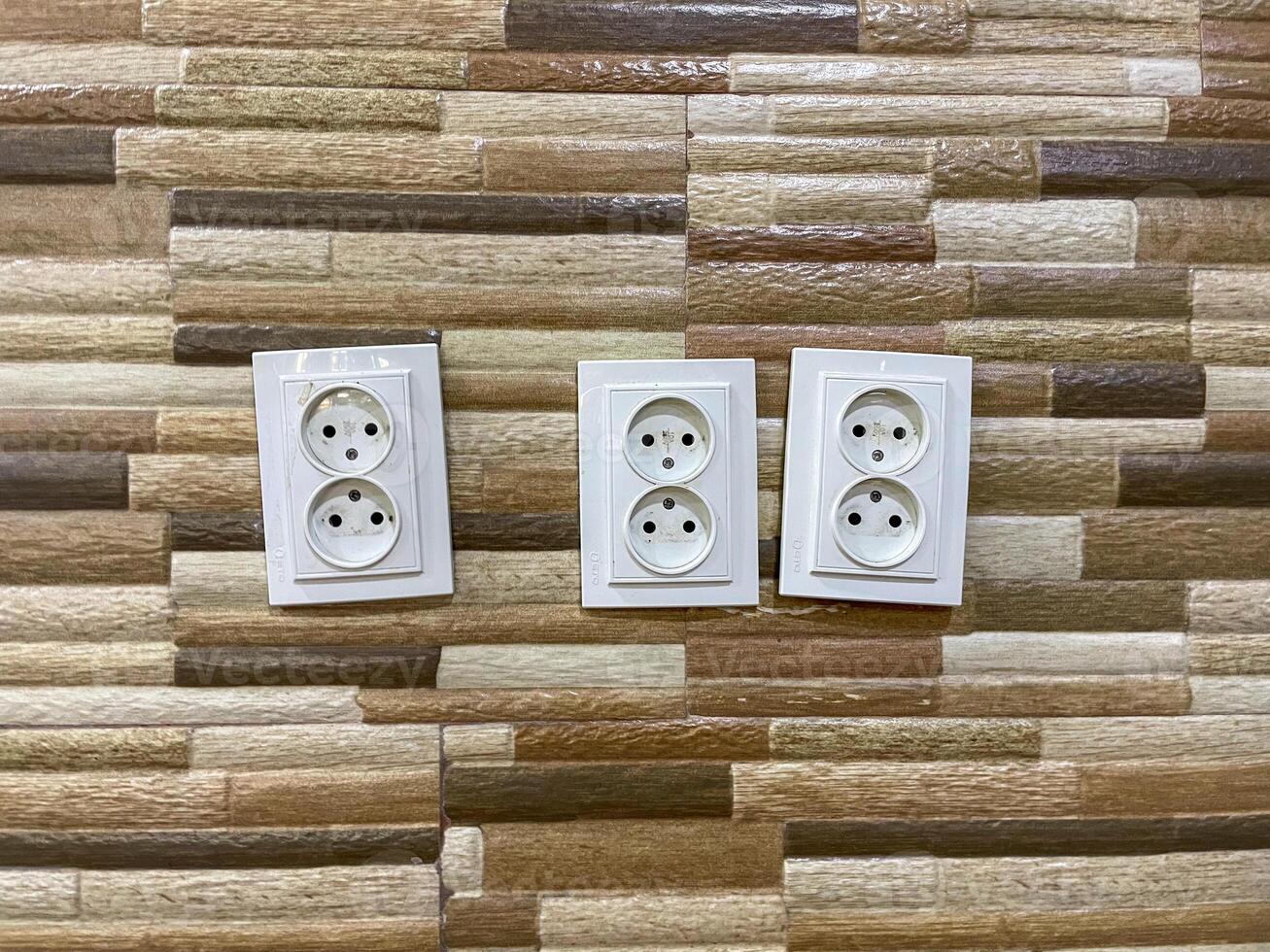 Three white double electrical outlets are mounted on a wall with a textured, multicolored tile pattern. The outlets are aligned horizontally and each has two sockets. photo
