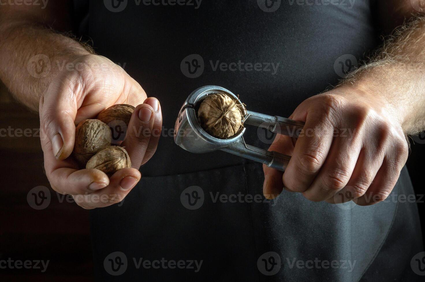 In a cozy kitchen, a person holds a handful of walnuts while using a nutcracker to split one open. The warm light highlights the earthy textures of the wood and nuts, creating a homey atmosphere photo