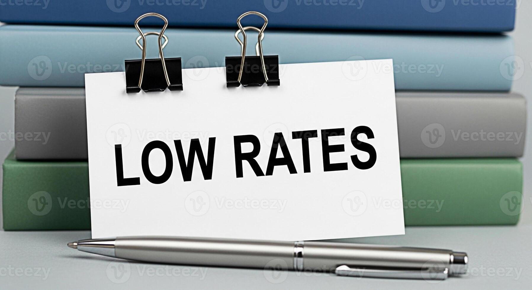 A crisp white card displaying Low Rates is clipped to a stack of colorful books accompanied by a sleek silver pen on a neutral surface conveying a message of affordability and value photo