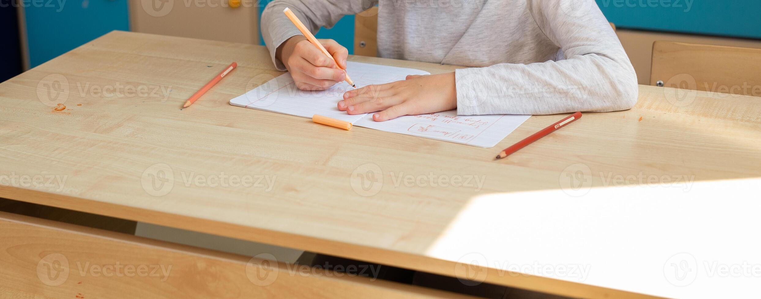 Child drawing in notebook with orange pencil during classroom lesson, banner with copy space. Creativity, imagination, and focus on self-expression through learning. photo