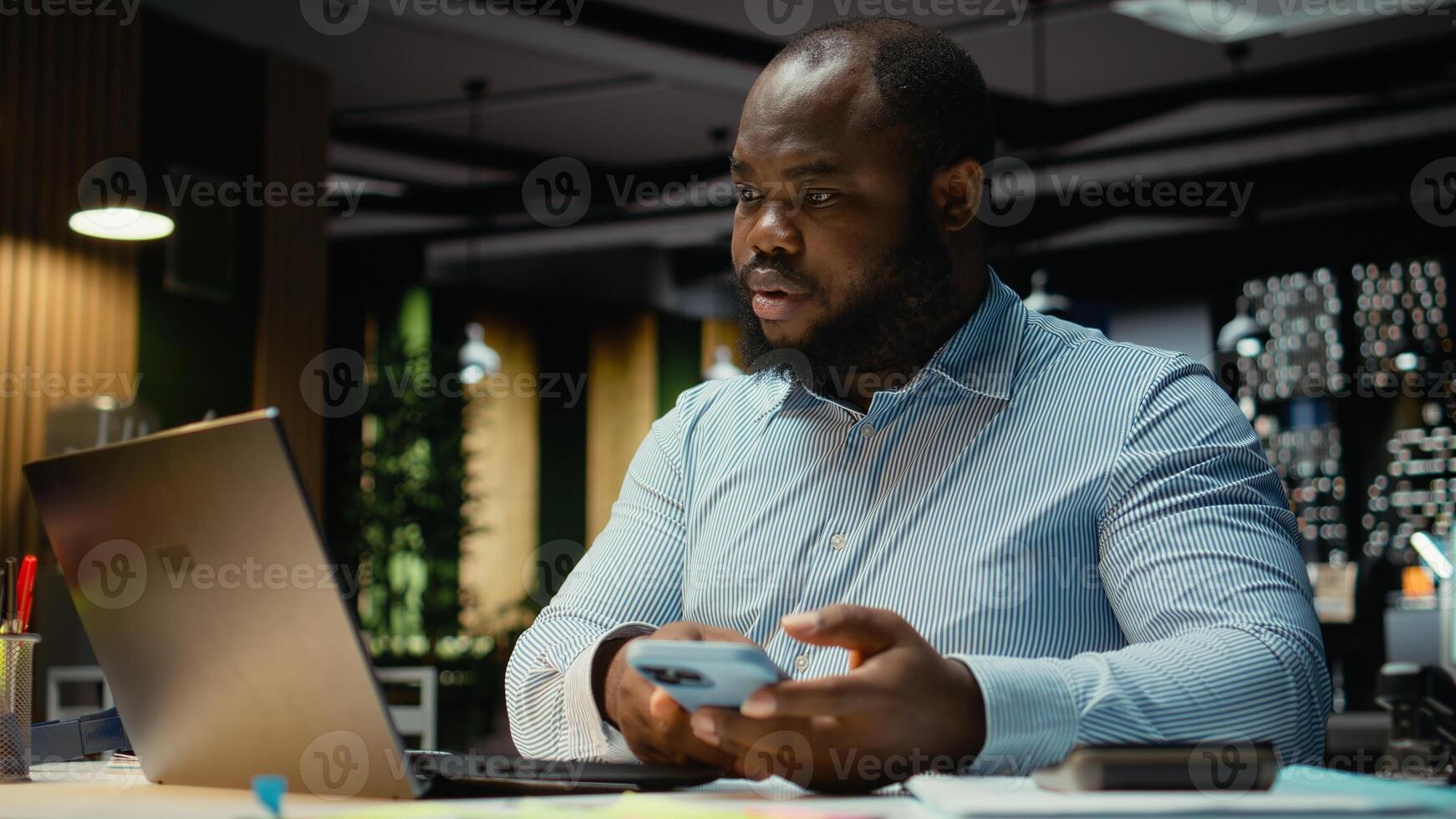 Black office employee checking a notification connected on socials while he does overtime at midnight, checking smartphone at work after hours. Young man reading text messages. photo