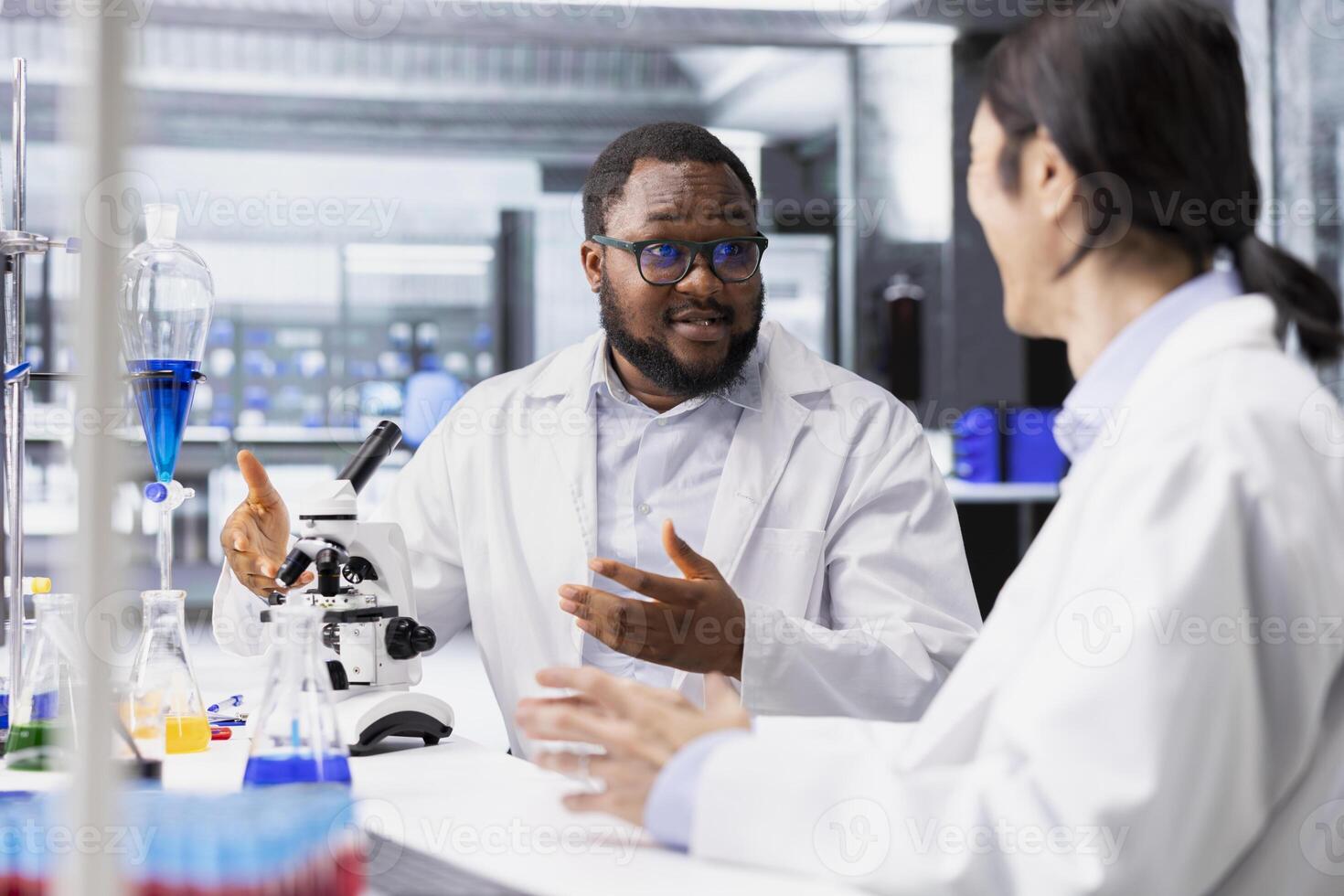Multiethnic team of microbiologists in laboratory inspecting specimen slide for microscopy study using optical device. Multiracial lab coworkers reviewing cytology slide for clinical testing procedure photo