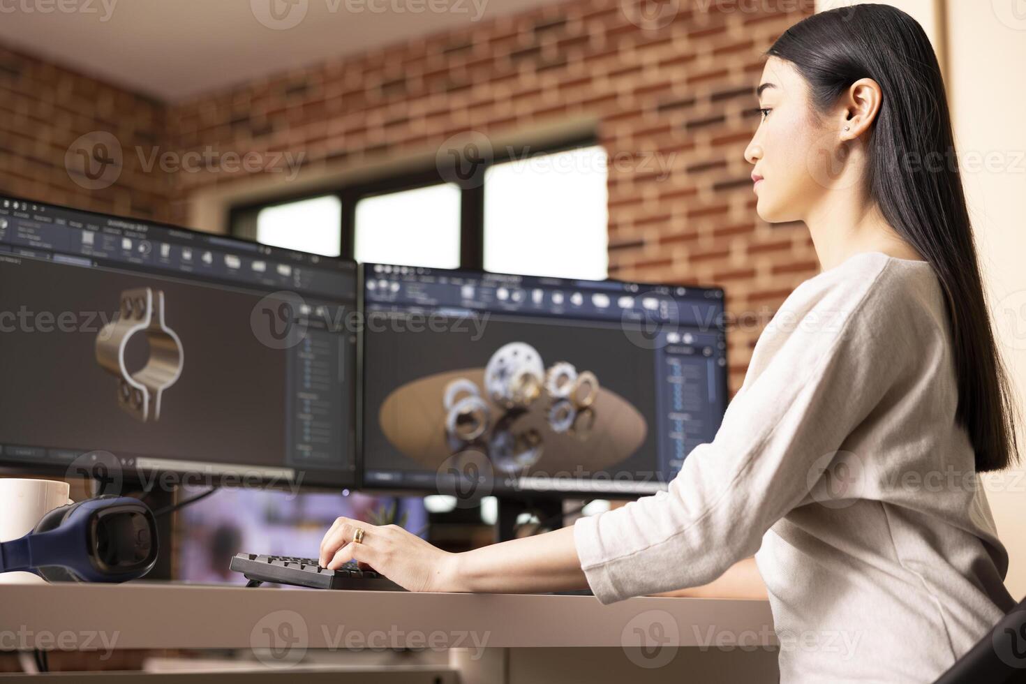 Focused female remote worker designing metal components using CAD software on desktop monitors. Industrial product asian developer analyzing 3D models on computer in brick wall home. photo