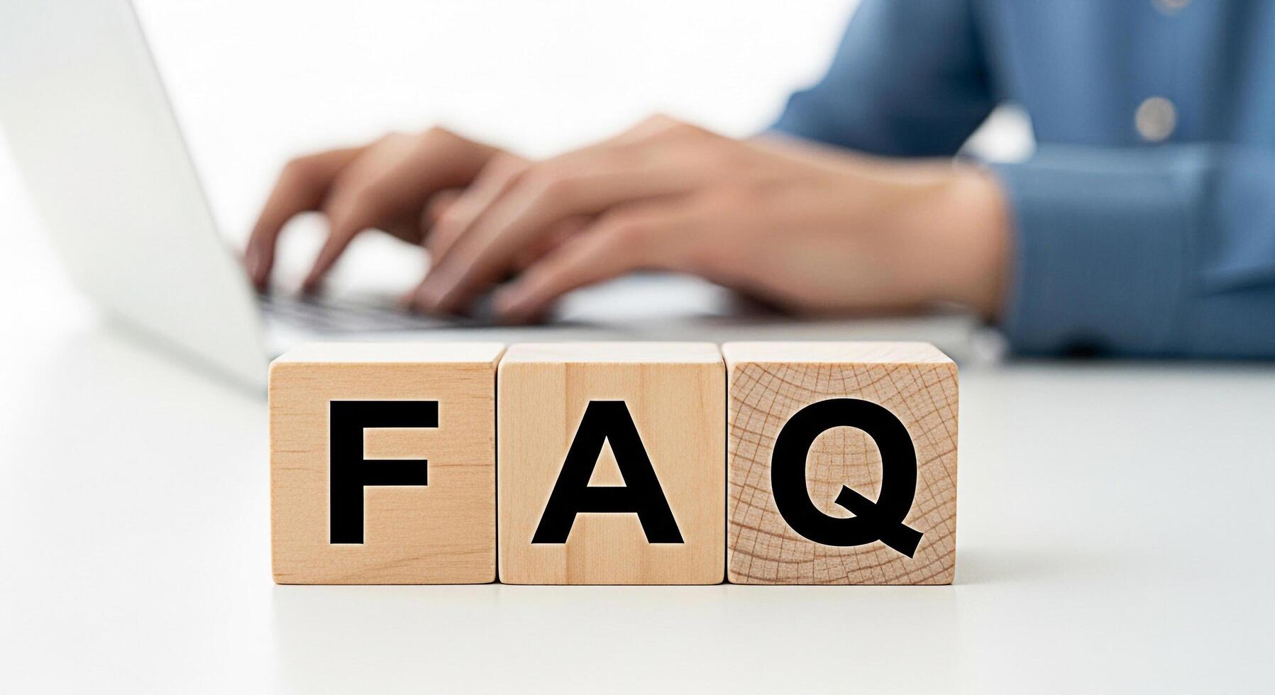 Person typing on a laptop with wooden blocks spelling FAQ on a white desk representing frequently asked questions and providing helpful information in a clean professional setting photo