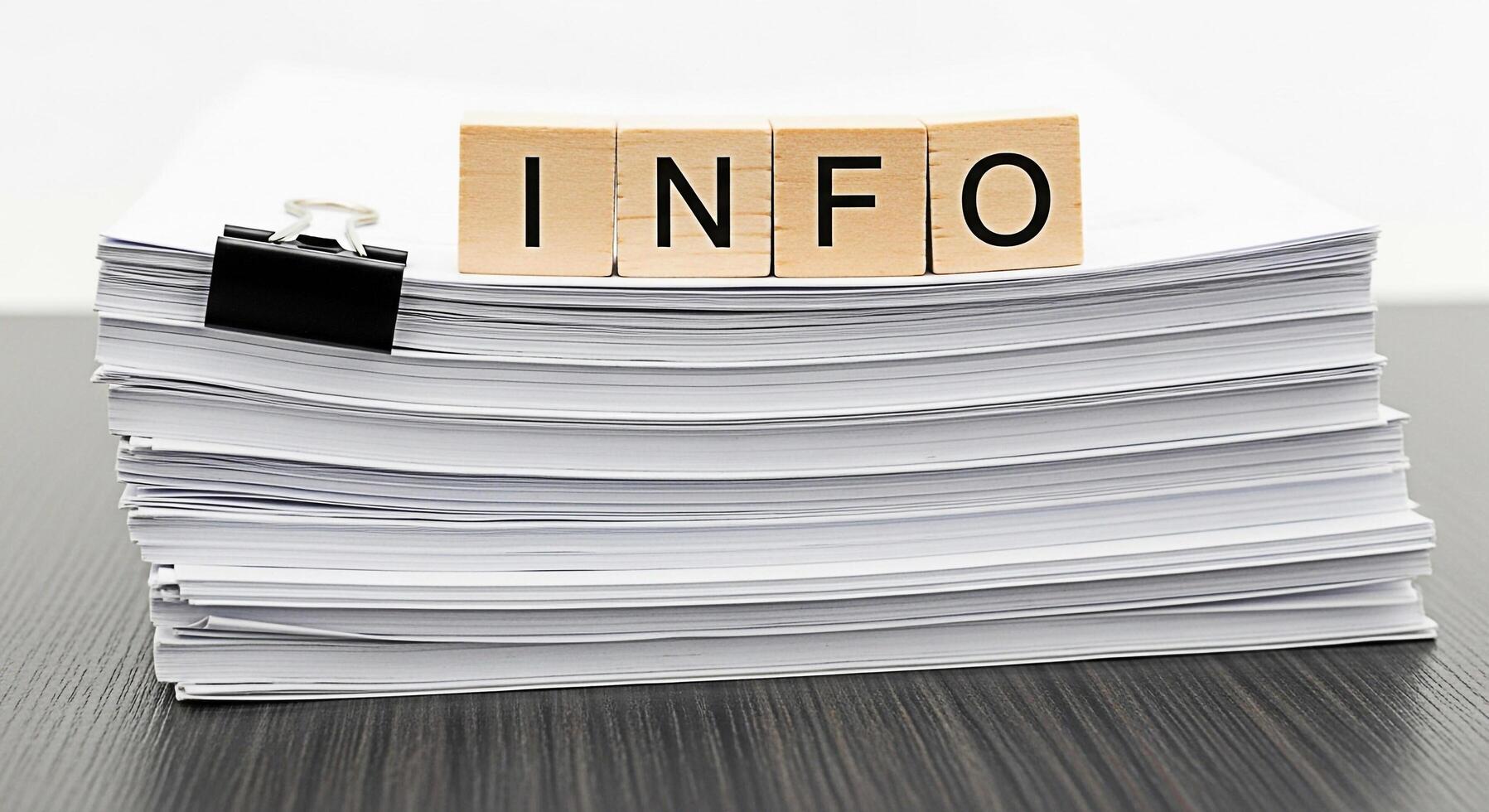 Stack of white papers with a black clip on a dark wooden desk displaying the word INFO spelled out in wooden blocks representing information overload and the importance of data management in business photo