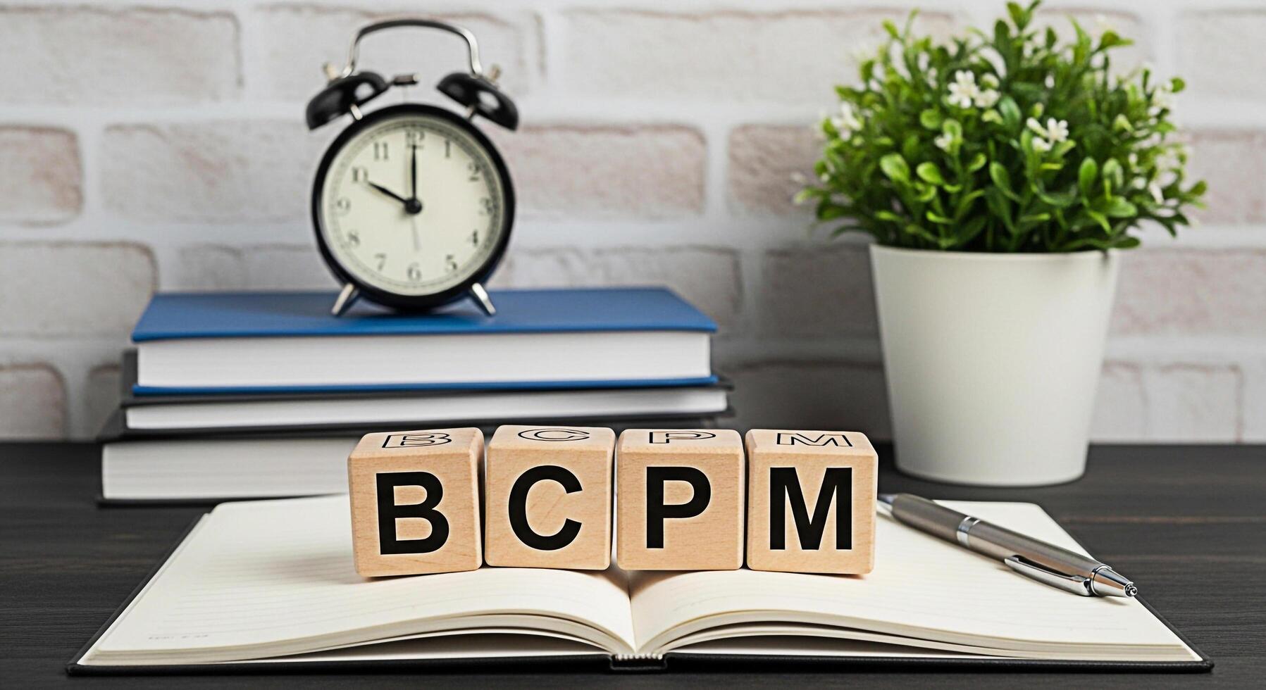 Wooden blocks spelling BCPM on an open notebook with a pen books alarm clock and plant against a brick wall symbolizing business continuity planning and management in a modern office environment photo
