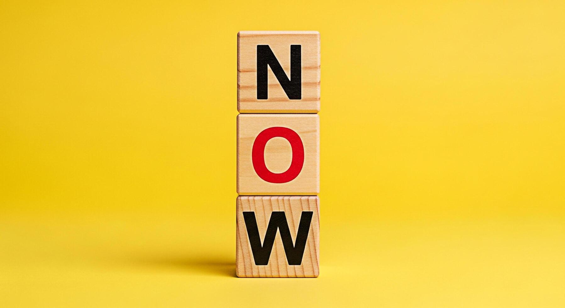 Wooden blocks spelling NOW stacked against a vibrant yellow background symbolizing urgency and immediate action creating a sense of motivation and the importance of seizing the moment inspiring prompt photo
