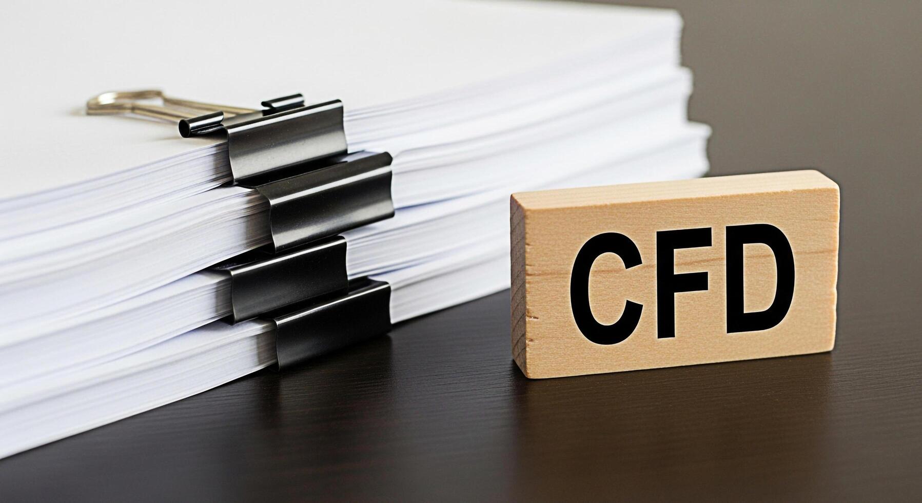 Stack of documents secured with binder clips next to a wooden block displaying CFD on a dark wooden desk representing contract for difference and financial analysis in a professional setting photo