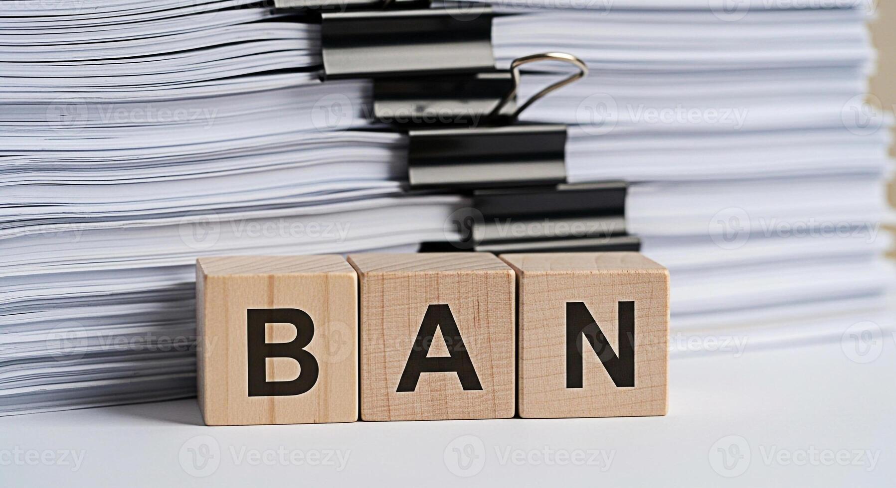 Wooden blocks spelling BAN stand defiantly against a towering stack of documents on a clean white surface symbolizing restriction and the urgent need to control excessive paperwork in a modern office photo