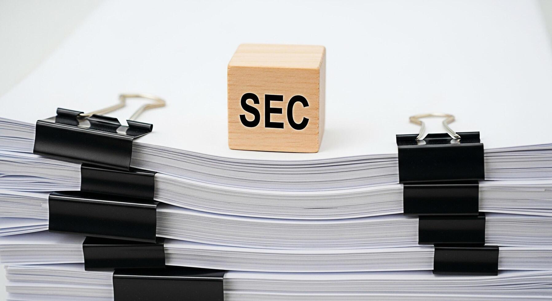 Wooden block displaying SEC sitting atop a stack of documents secured with binder clips in a bright minimalist office setting symbolizing regulatory compliance and financial oversight with a sense of photo