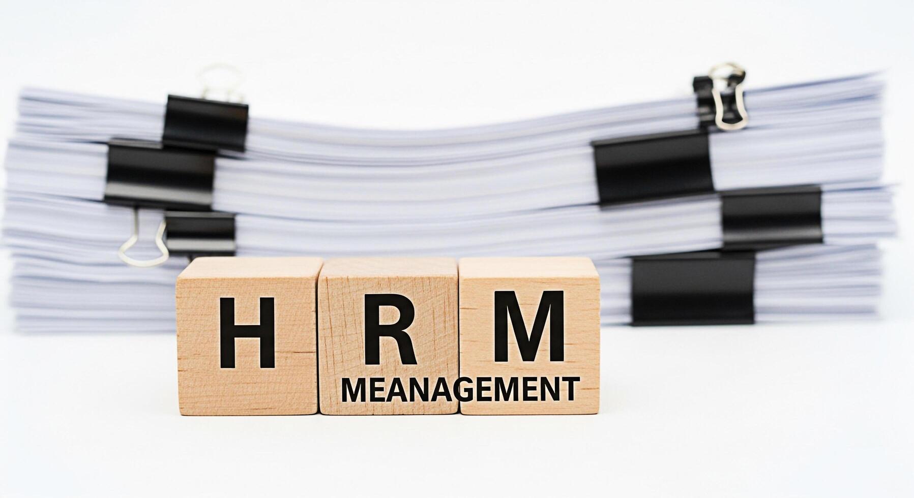 Wooden blocks spelling HRM Management in front of stacked documents on a white background representing human resources management paperwork overload and the importance of efficient HR processes photo