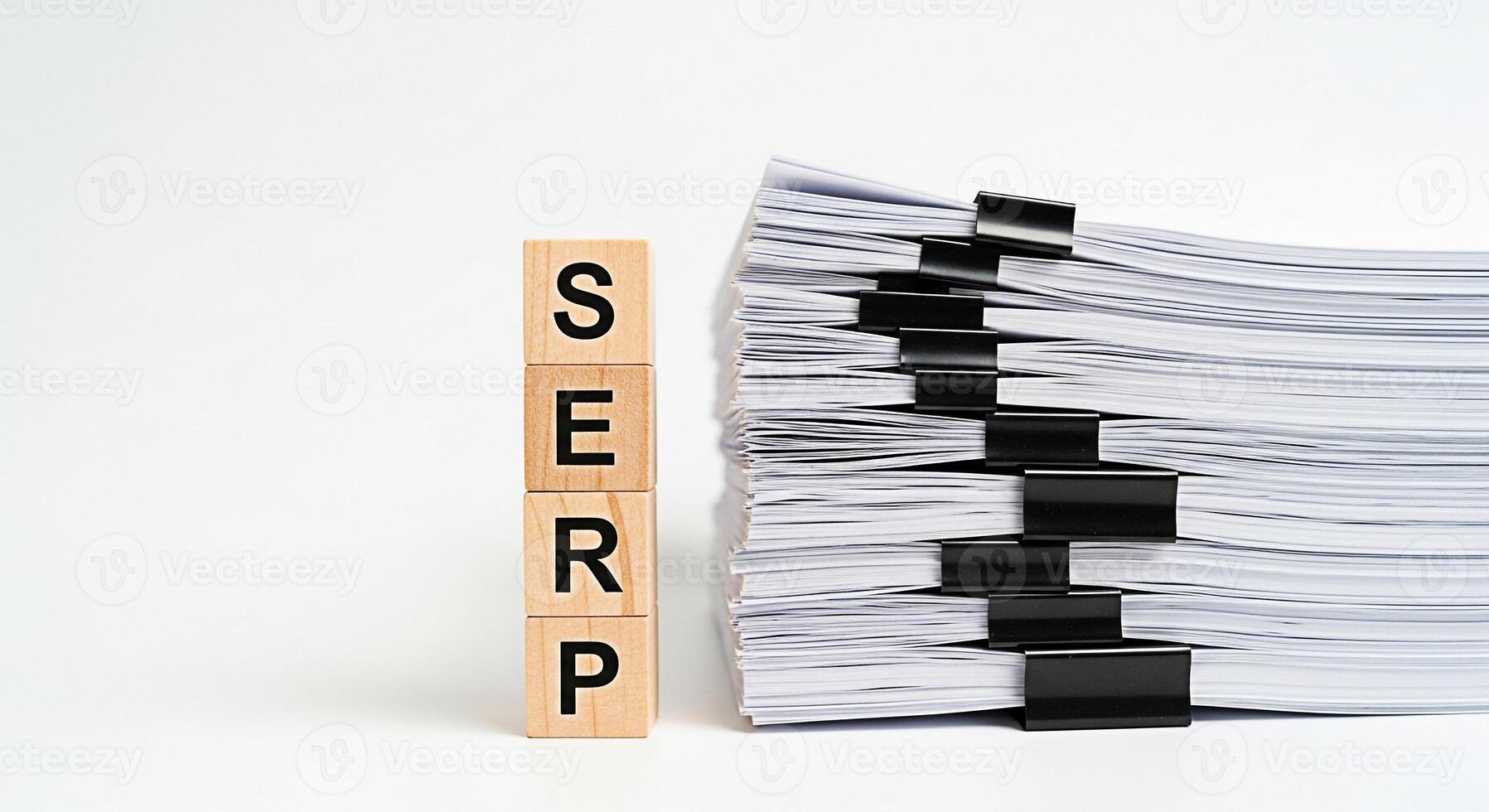 Wooden blocks spelling SERP stand next to a large stack of documents secured with binder clips on a white surface representing search engine optimization and the importance of content creation photo