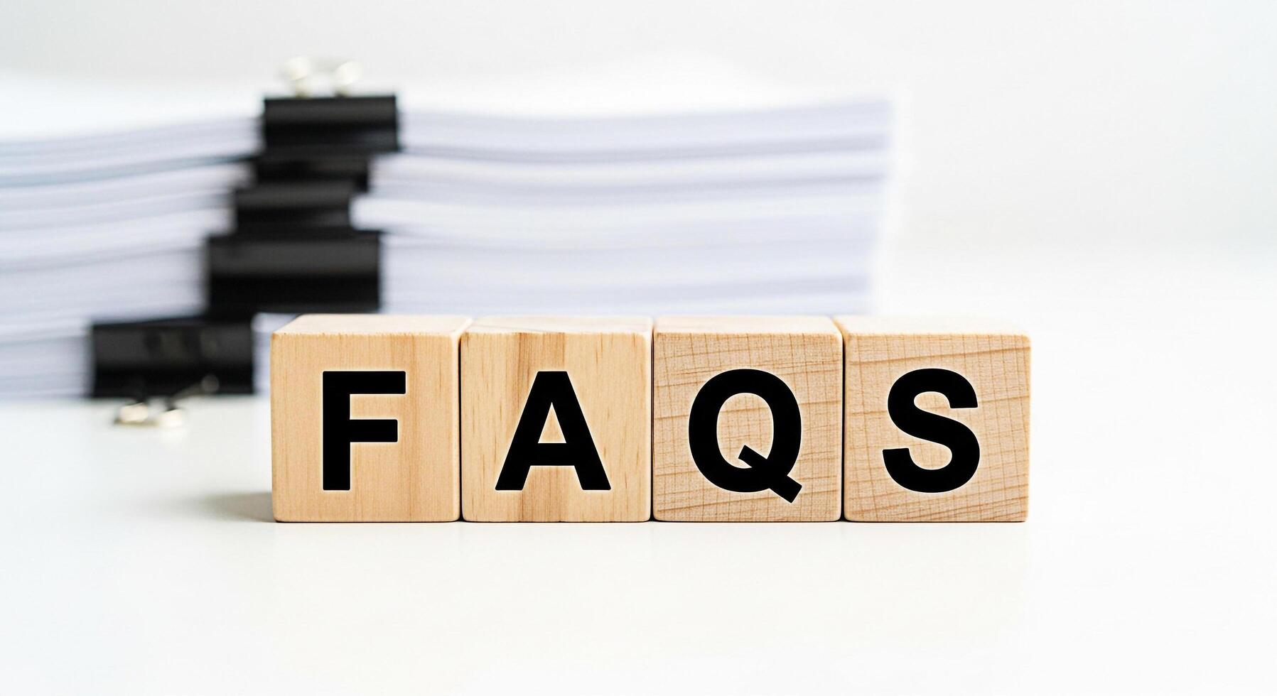 Wooden blocks displaying FAQs on a white desk with a stack of papers representing frequently asked questions and providing answers in a clear and concise manner for customer support and information photo