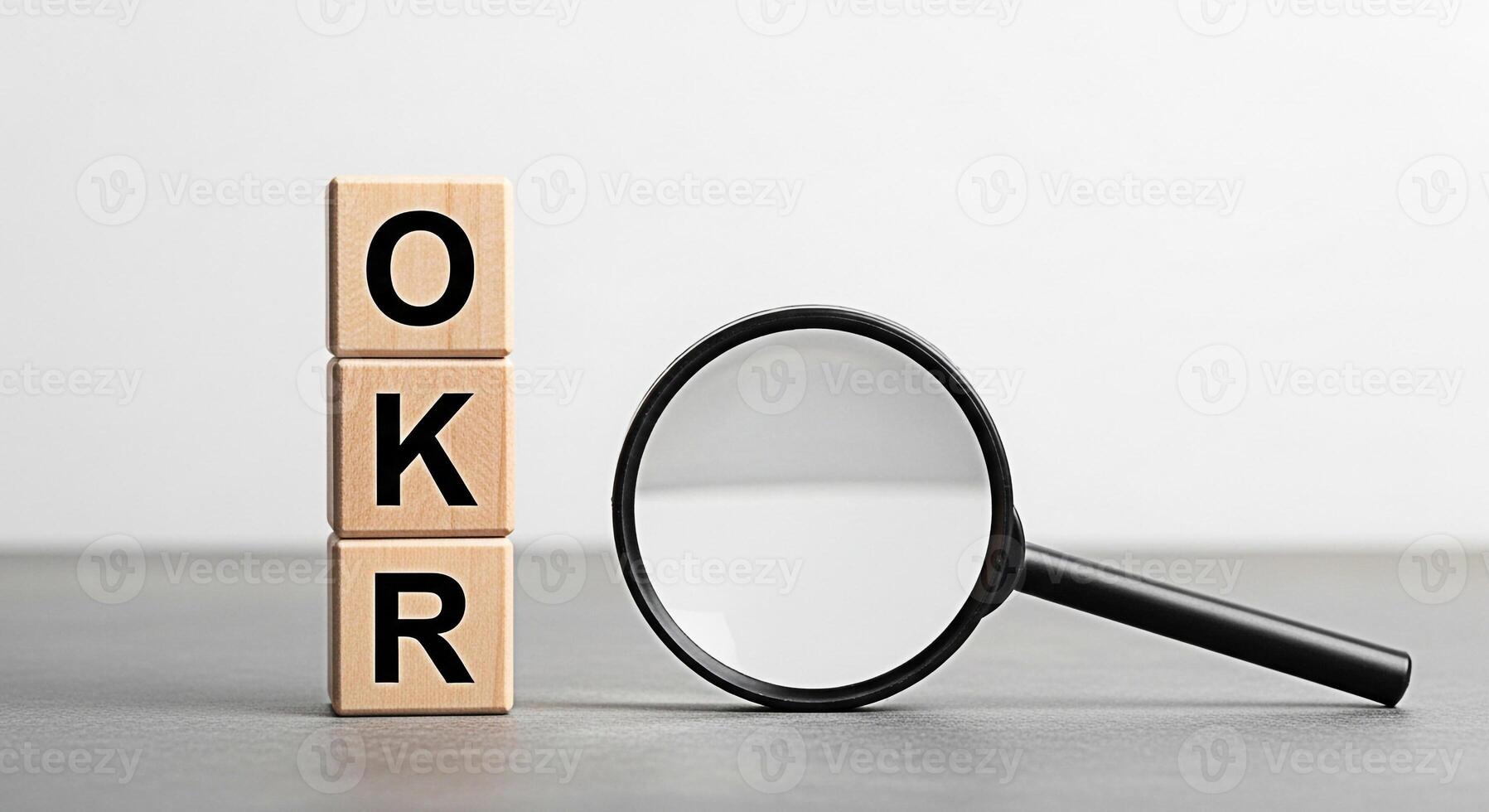 Wooden blocks displaying OKR acronym under scrutiny with a magnifying glass on a gray surface symbolizing the importance of setting and achieving objectives and key results in a business environment photo