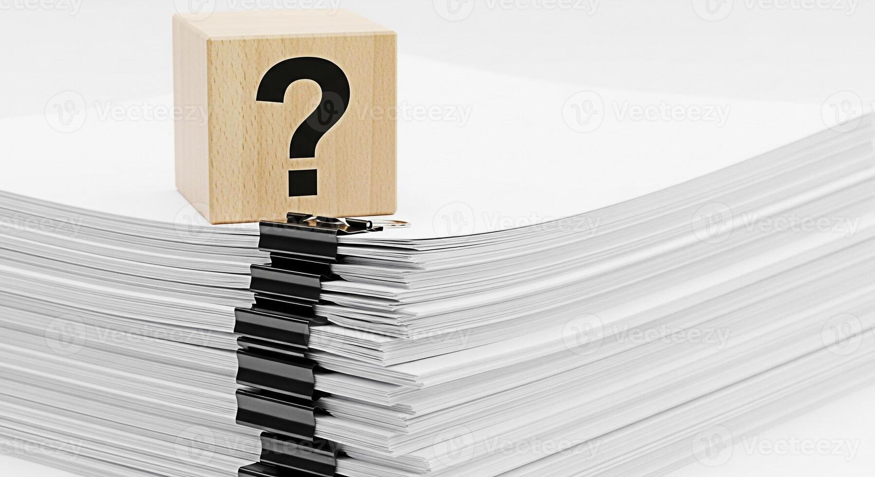 Wooden cube with question mark sitting atop a stack of white paper held together with binder clips symbolizing uncertainty and the need for answers in business and document management photo