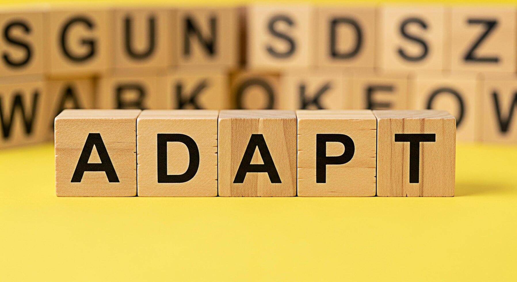 Wooden blocks spelling ADAPT on a yellow surface with blurred letter blocks in the background symbolizing the need to adjust and change in a dynamic environment conveying a message of resilience and f photo