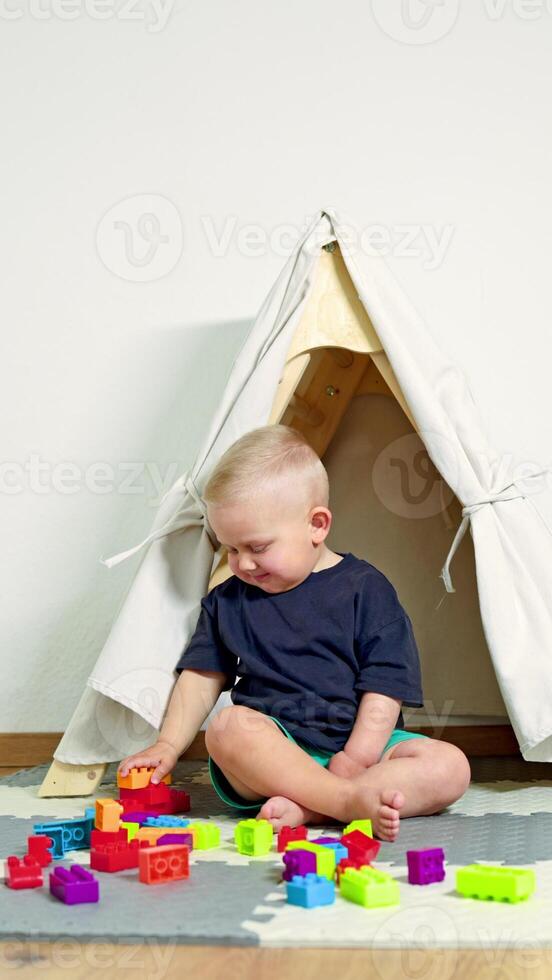 Toddler boy sits home on a rug and builds a tower from a construction set. Self-development of a child, development of fine motor skills of hands in children. Child plays with a construction set photo