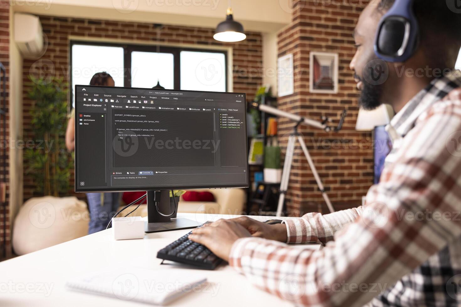 Remote male programmer sits at desk with headphones, coding and debugging scripts on computer. African american man listens to music and works on desktop pc, creating web application from home office. photo