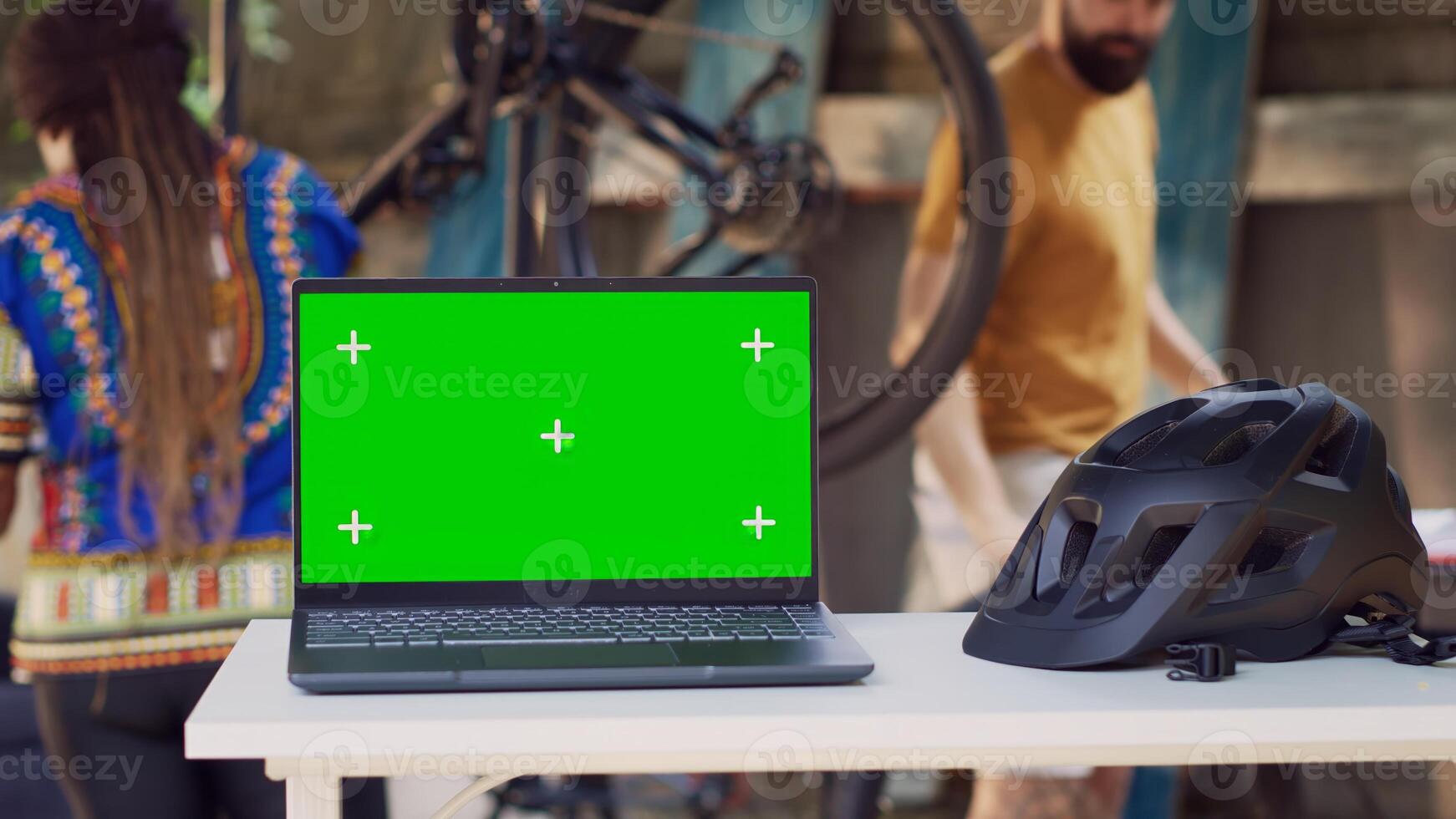 Close-up of laptop with greenscreen display placed near helmet while interracial couple performs bike maintenance in background. In yard minicomputer is on table showing isolated chromakey template. photo