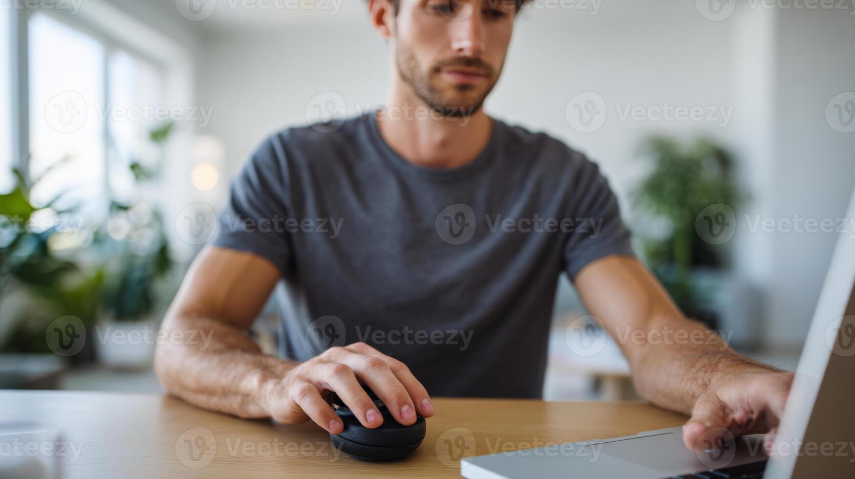 A focused young Caucasian man using a laptop and computer mouse in a bright, modern workspace filled with greenery. photo