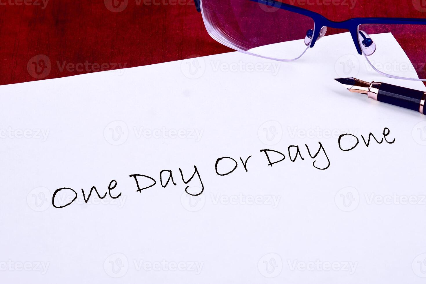 A handwritten phrase, One Day Or Day One, rests on white paper next to a fountain pen and reading glasses, set against a red background, conveying a message of ambition and choices photo