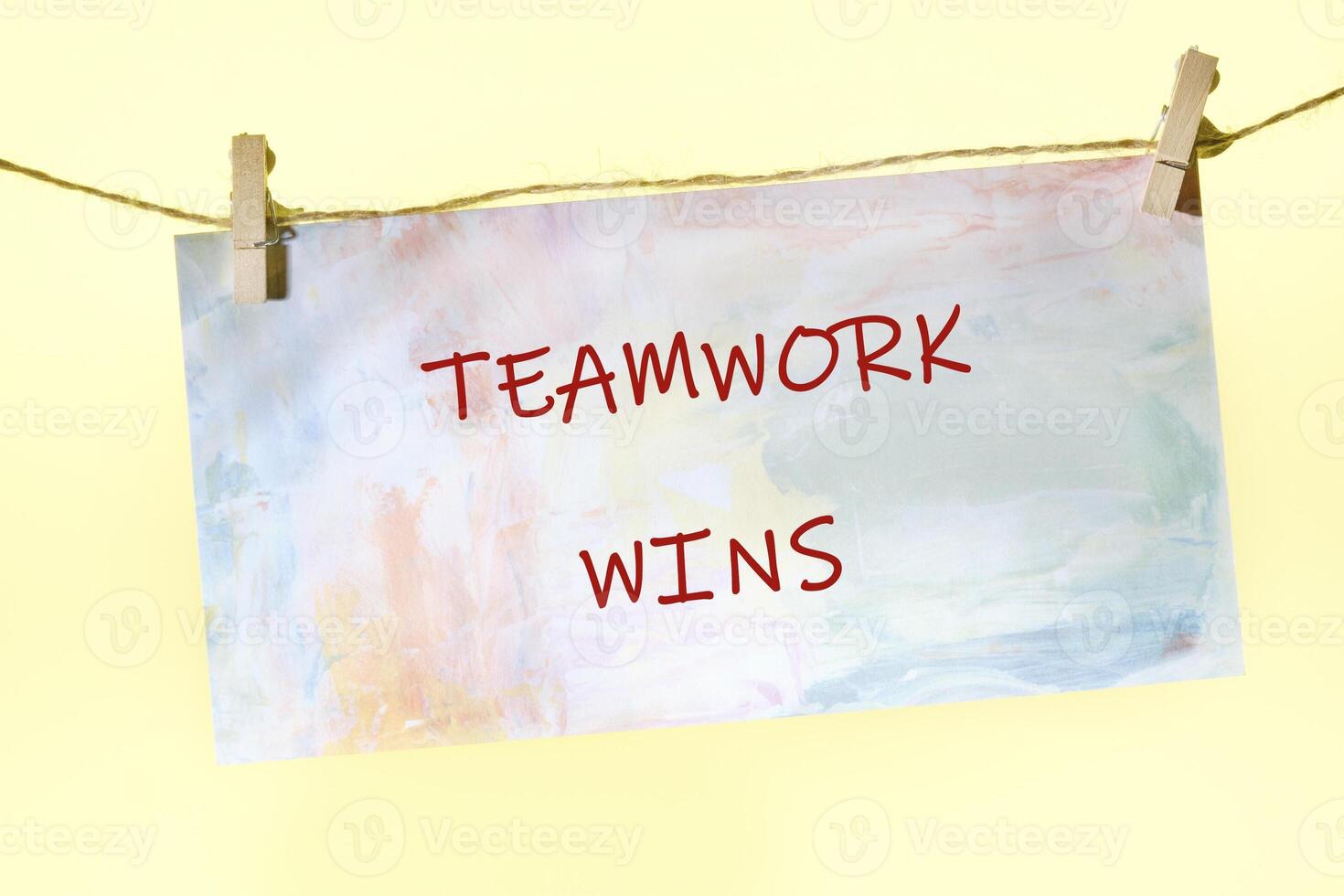 A sign with the phrase TEAMWORK WINS hangs from a clothesline using clothespins. The background is a simple yellow, adding to the optimistic message photo