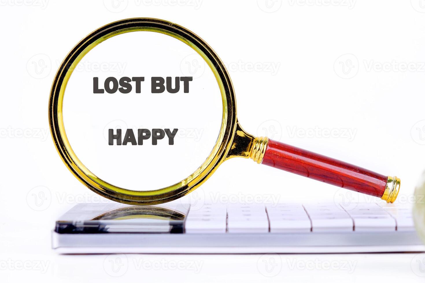 A magnifying glass with a red handle and gold rim shows the words lost but happy. It rests on top of a white computer keyboard. The background is plain white photo