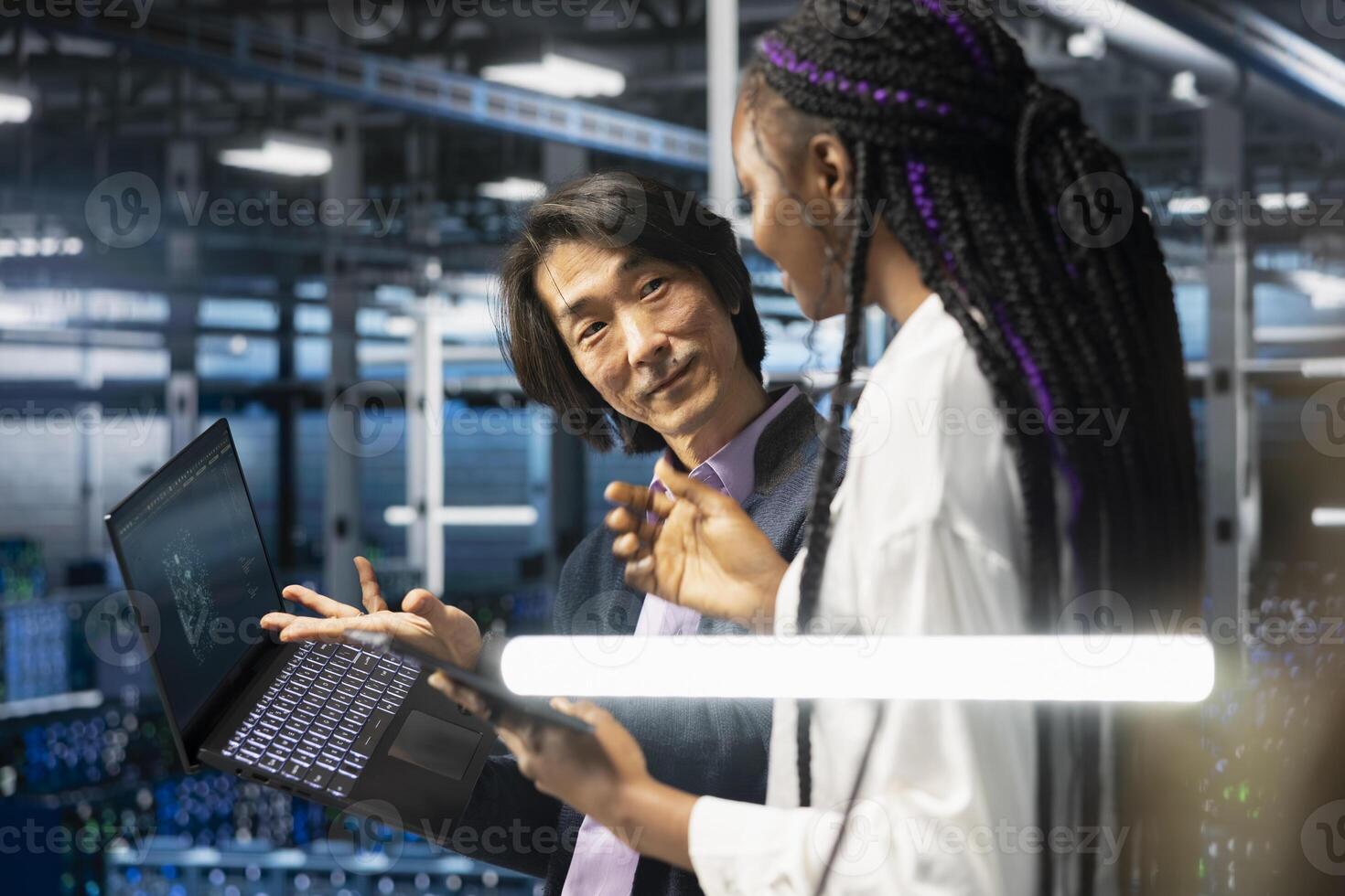 Multiethnic coworkers in diverse data center installing upgraded server racks with AI. African american woman and Asian senior programmer in server room upgrading gear using artificial intelligence photo