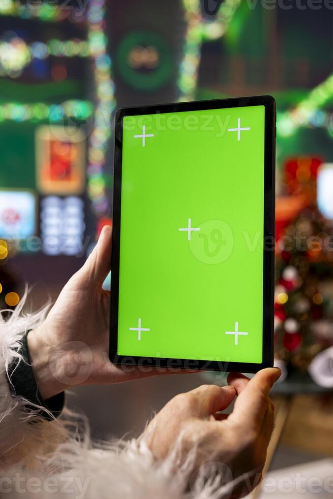 Close up of Santa Claus using mockup tablet in North Pole workshop to read letters from kids. Saint Nicholas answering online messages from children using green screen touchscreen device photo