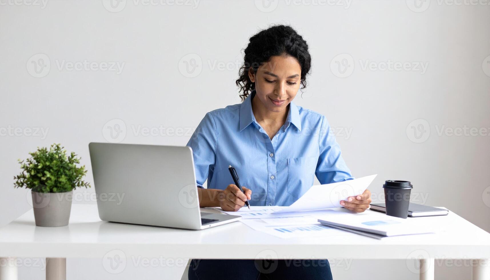 Woman is sitting at desk, focused on her work. She is reviewing documents and taking notes while using laptop. small plant and coffee cup are on table, creating productive atmosphere photo