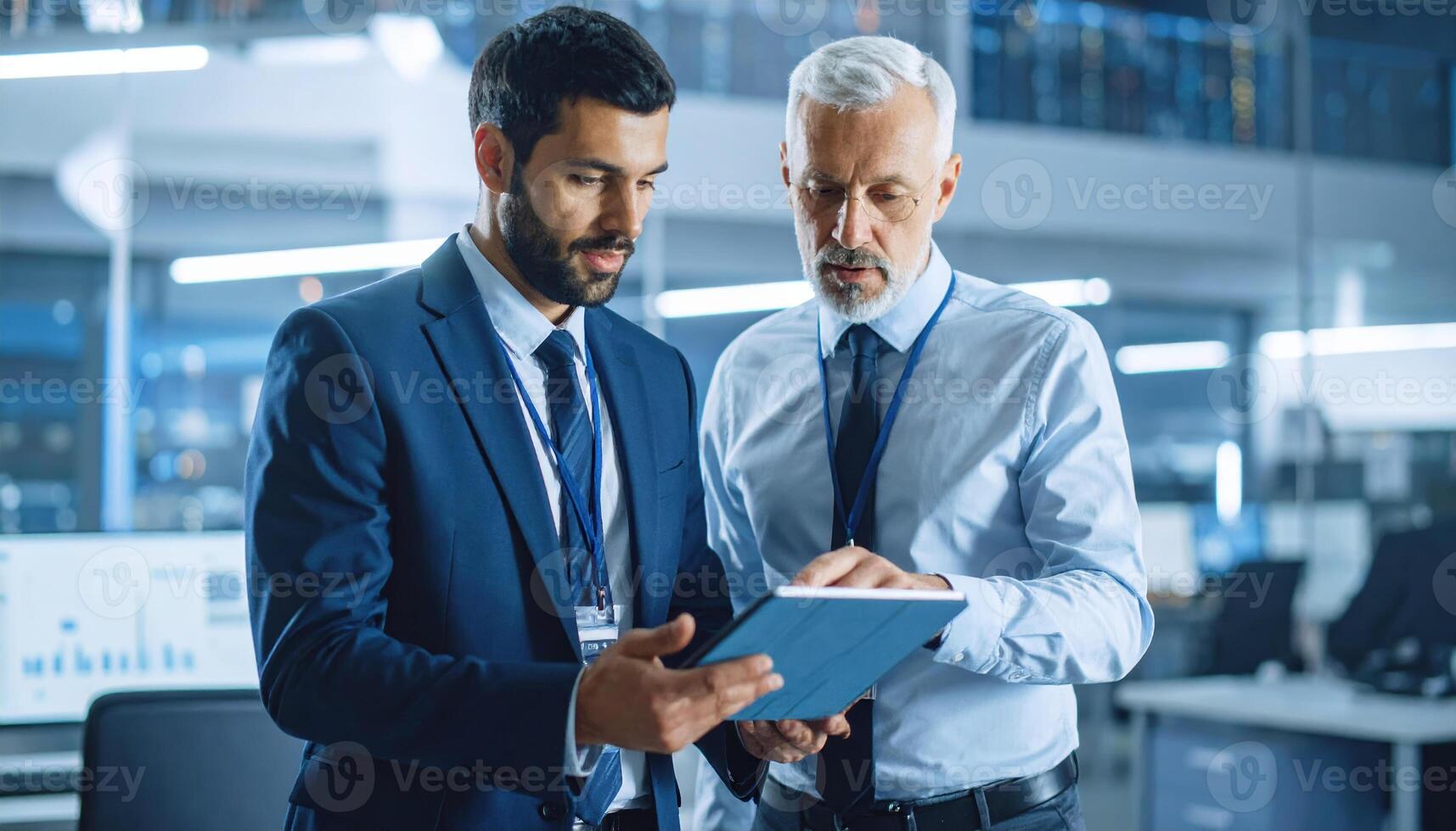 Two businessmen in formal suit and shirt discussing data on digital tablet in modern office environment with technology and charts, showing focused and professional mood photo