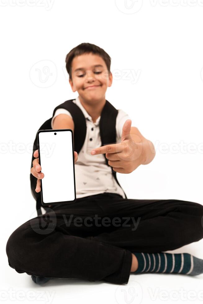 Young boy with backpack sits cross-legged, holding and pointing at an isolated blank smartphone on white background. Suitable for technology, education, or advertising concepts requiring isolated device display. photo