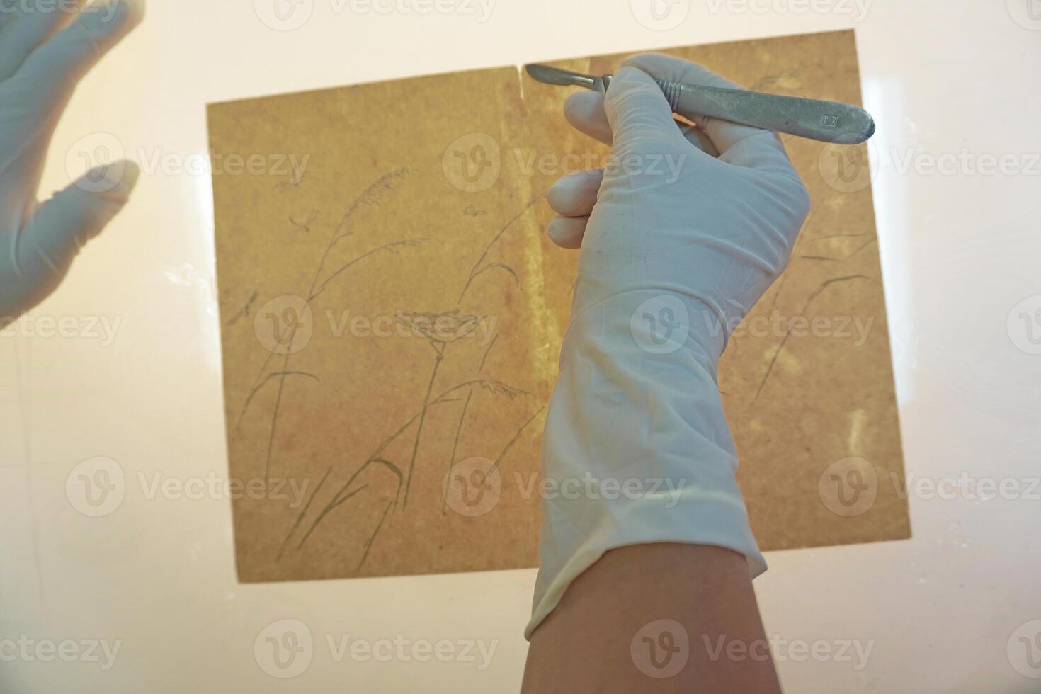 Gloved hands work with a scalpel on a light table. Processing of a drawing on a transparent sheet, archival restoration of the document. photo