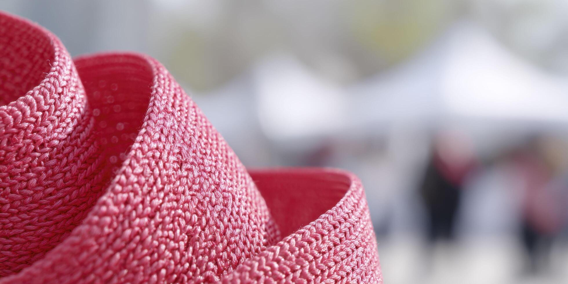 A macro view of red fabric woven into a ribbon pattern, highlighting its tight threads and soft sheen under daylight, with a blurred World AIDS Day booth and crowd in the background. photo