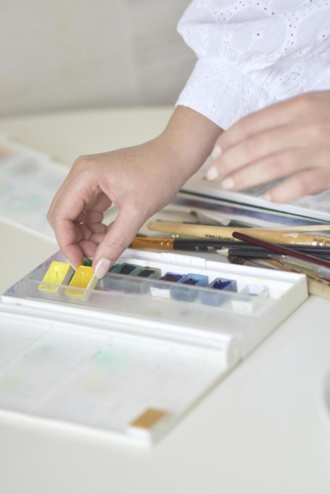 A close-up of a hand picking a yellow watercolor block from a paint set, with brushes and a palette visible. Creative, colorful, and cozy mood suited for art, hobbies, and craft projects. photo