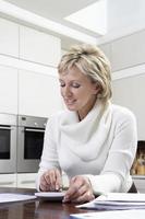 Woman Calculating Domestic Bills With Calculator In Kitchen photo