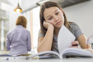 Bored girl studying at table with mother standing in background photo