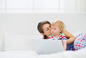 joven madre besando a su bebé moderno trabajando en la computadora portátil foto