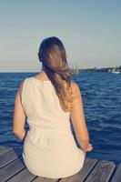 Woman in white dress sitting on dock, looking toward ocean photo
