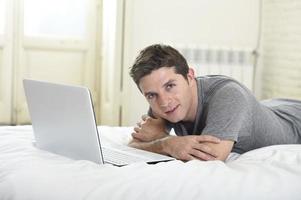 joven atractivo hombre acostado en la cama disfrutando de la computadora de redes sociales foto