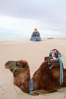 Caucasian man sitting on sand dune in desert with camel photo