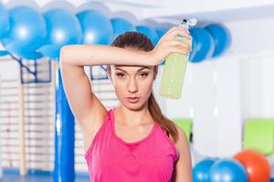Young girl drinking isotonic drink, gym. She is wiping sweat photo