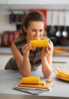 Retrato de joven ama de casa feliz comiendo maíz hervido en la cocina