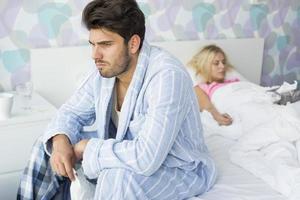 Sick man sitting on bed with woman sleeping