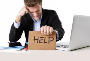 businessman working in stress at computer holding help cardboard sign