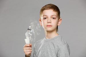 Unhappy boy holding inhalator mask releasing smoke on grey background photo