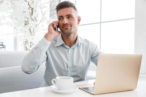 Apuesto hombre de negocios trabajando con ordenador portátil y hablando por teléfono en el café